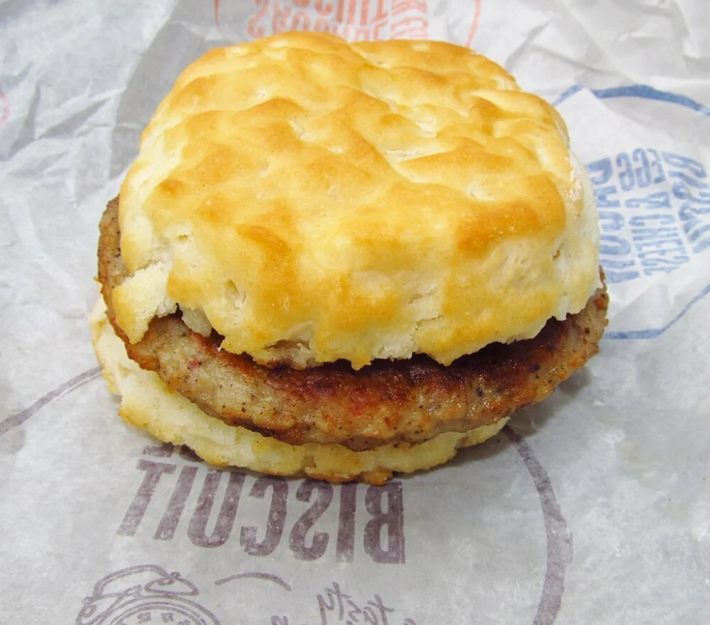 A close-up of a sausage biscuit with a golden, flaky biscuit and a seasoned sausage patty on branded fast-food packaging.